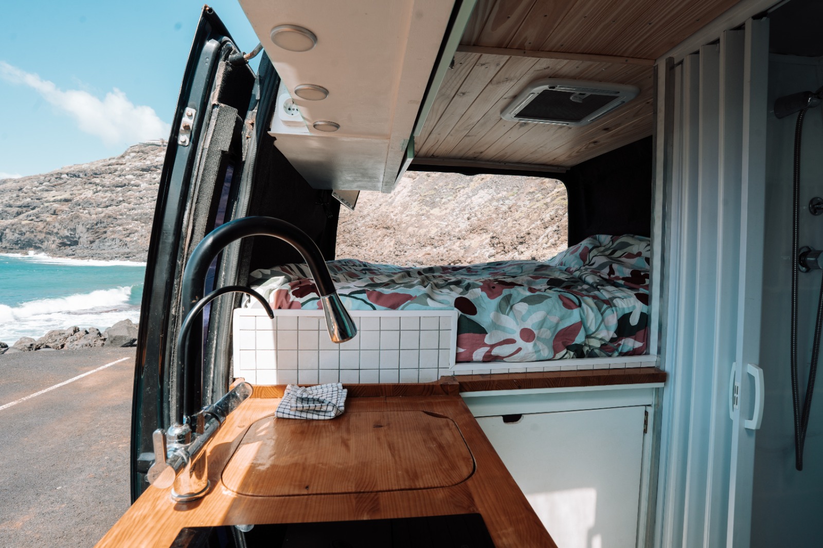 Campervan interior in Tenerife with the side door open to a rocky beach, a wooden kitchen counter with sink and a made-up double bed